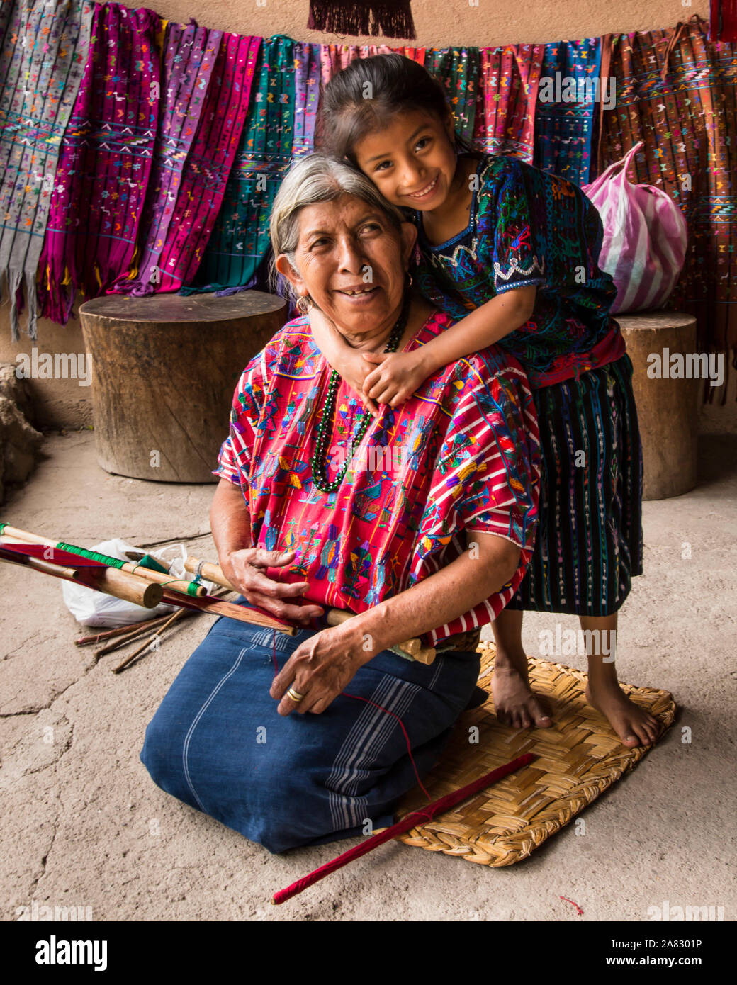 A young Guatemalan Mayan girl in traditional dress, hugs her grandmother, who is kneeling and weaving on a backstrap loom.   Santa Catarina Palopo, Gu Stock Photo