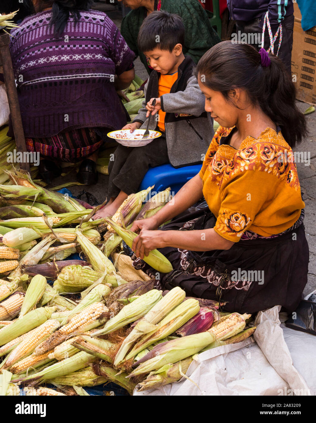 A woman husks corn at the market in Chichicastenango, Guatemala while her young son eats.  The boy appears to be about 4 years old. Stock Photo