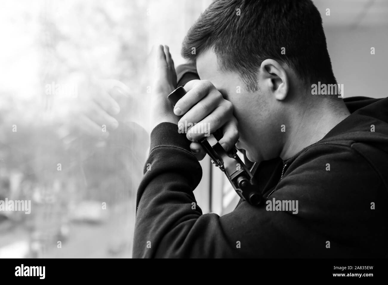 Black and white photo of young man with gun going to commit suicide near window Stock Photo