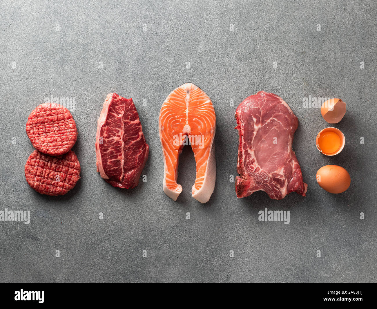 Carnivore or keto diet concept. Raw ingredients for zero carb or low carb diet - burger patties, ribeye, salmon steak, pork, egg on gray stone background.Top view or flat lay.Copy space top and bottom Stock Photo