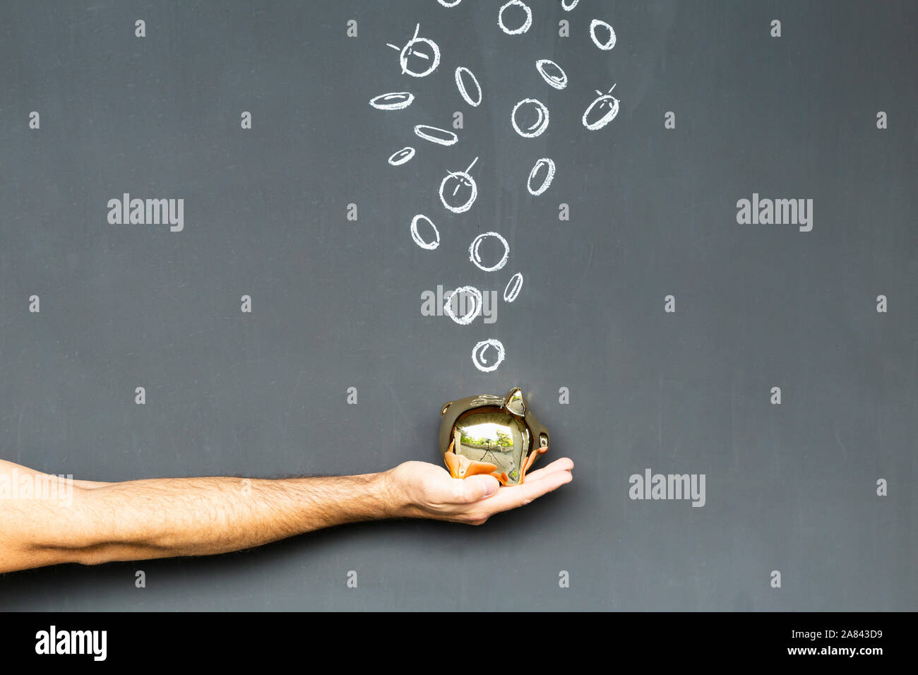 Concept of saving money with a gold colored piggy bank held in a hand in front of a blackboard with hand drawn coins Stock Photo