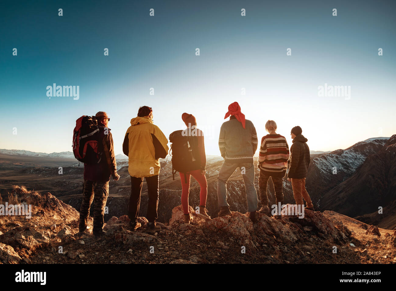 Group of six tourists backpackers or hikers stands against sunset light and mountains Stock Photo