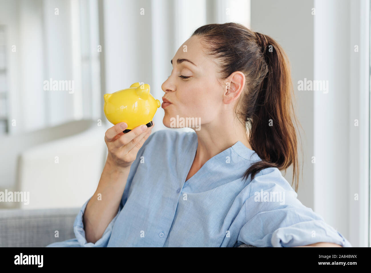 Successful young woman kissing a piggy bank as she relaxes on a sofa as she imagines how she is going to spend her nest egg Stock Photo