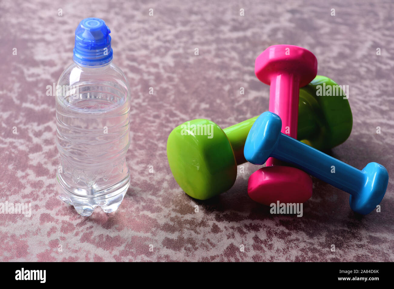 Dumbbells made of plastic near water bottle on burgundy texture background.  Barbells in green, pink and cyan color and water, close up. Shaping and fitness  equipment. Healthy shape and sport concept Stock