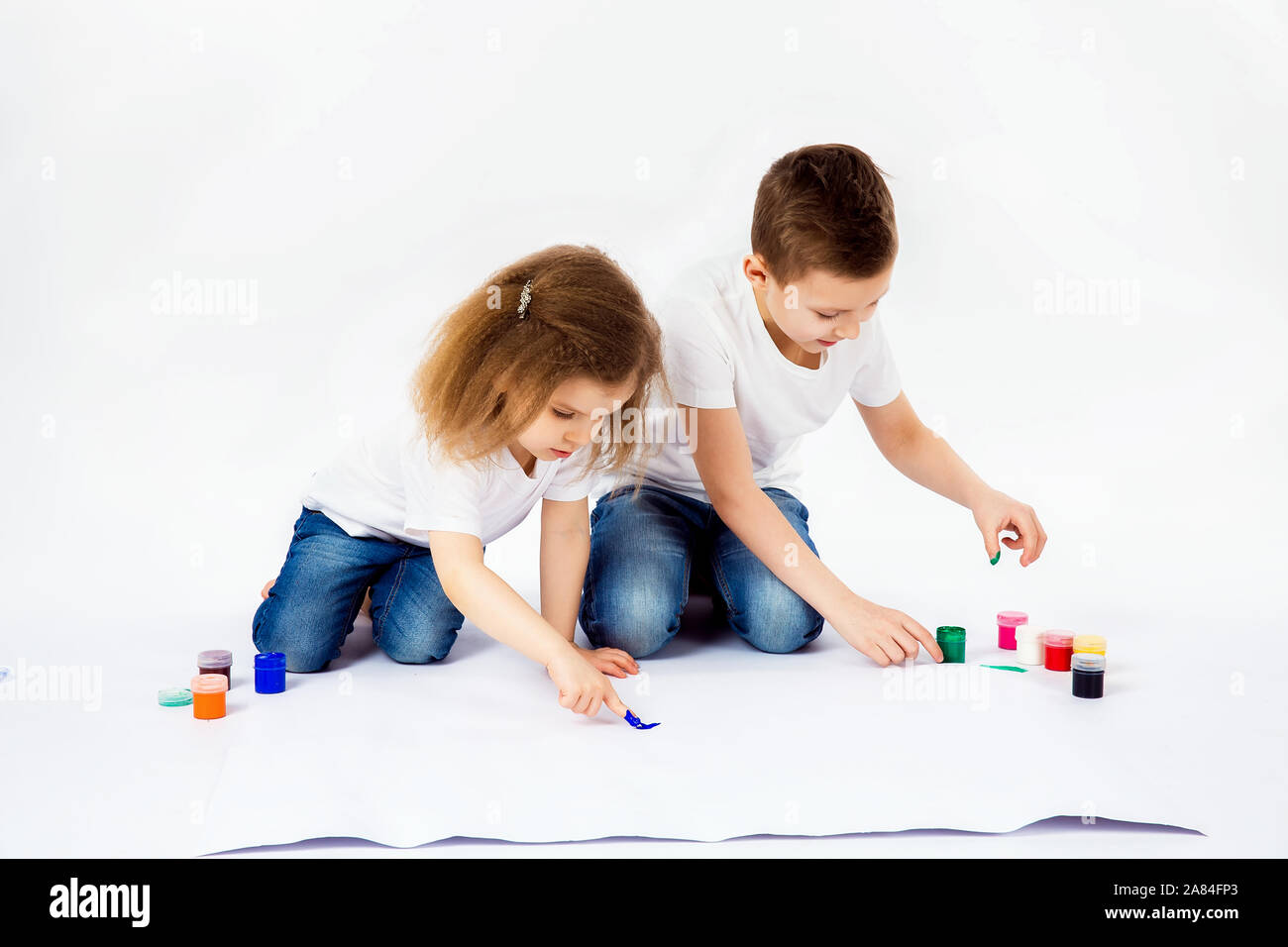 Two Pretty Child Friends Boy And Girl In White Shirts And Blue Jeans Trendy Hair Style Barefoot Drawing Pictures On White Sheet Of Paper By Paints Isolated On White Studio Shot Stock