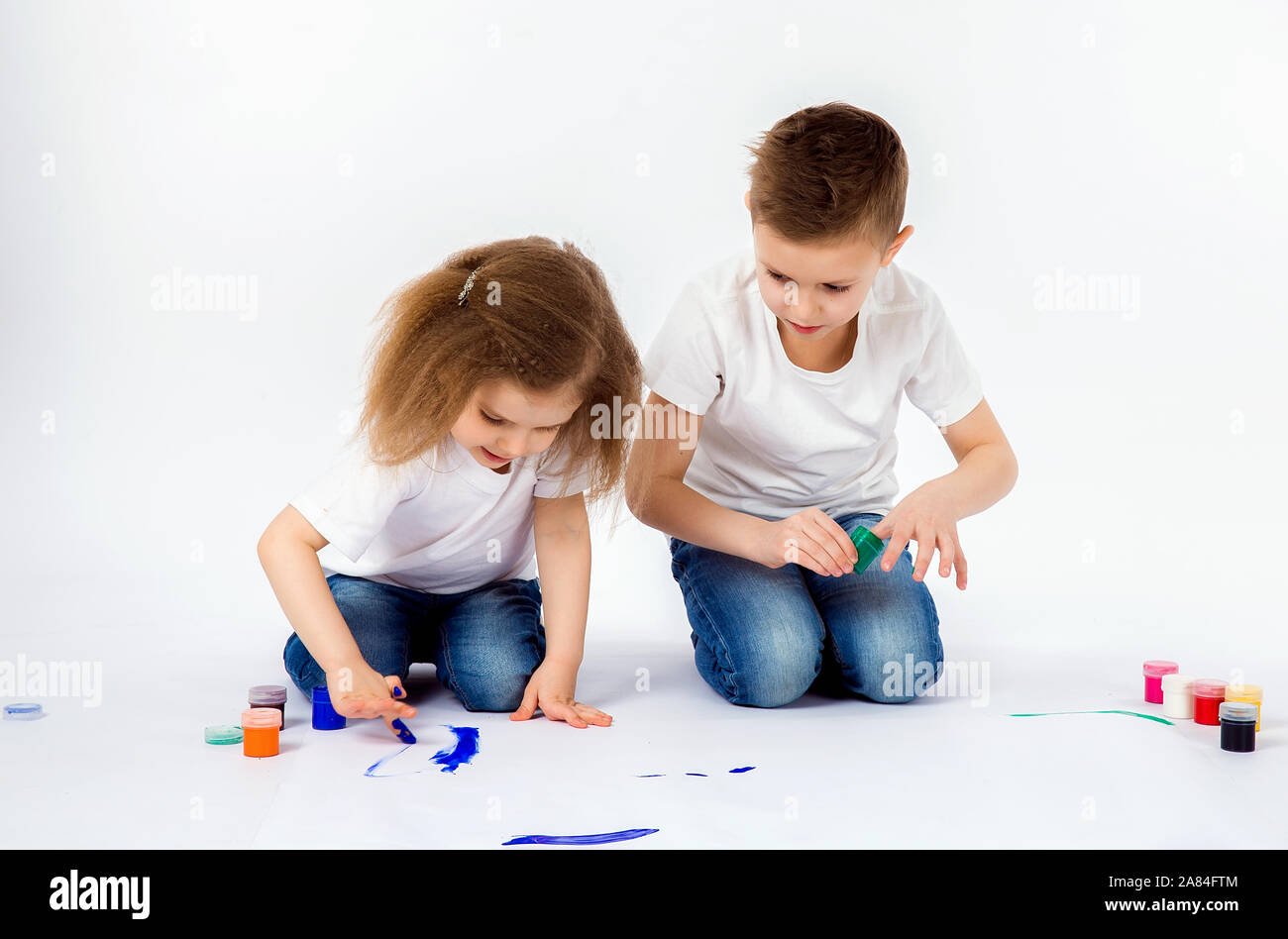 Two Pretty Child Friends Boy And Girl In White Shirts And Blue Jeans Trendy Hair Style Barefoot Drawing Pictures On White Sheet Of Paper By Paints Isolated On White Studio Shot Stock