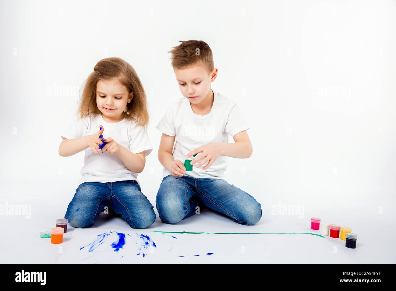 Two pretty child friends boy and girl in white shirts and blue jeans,  trendy hair style, barefoot, drawing pictures on white sheet of paper by  paints isolated on white. Studio shot Stock