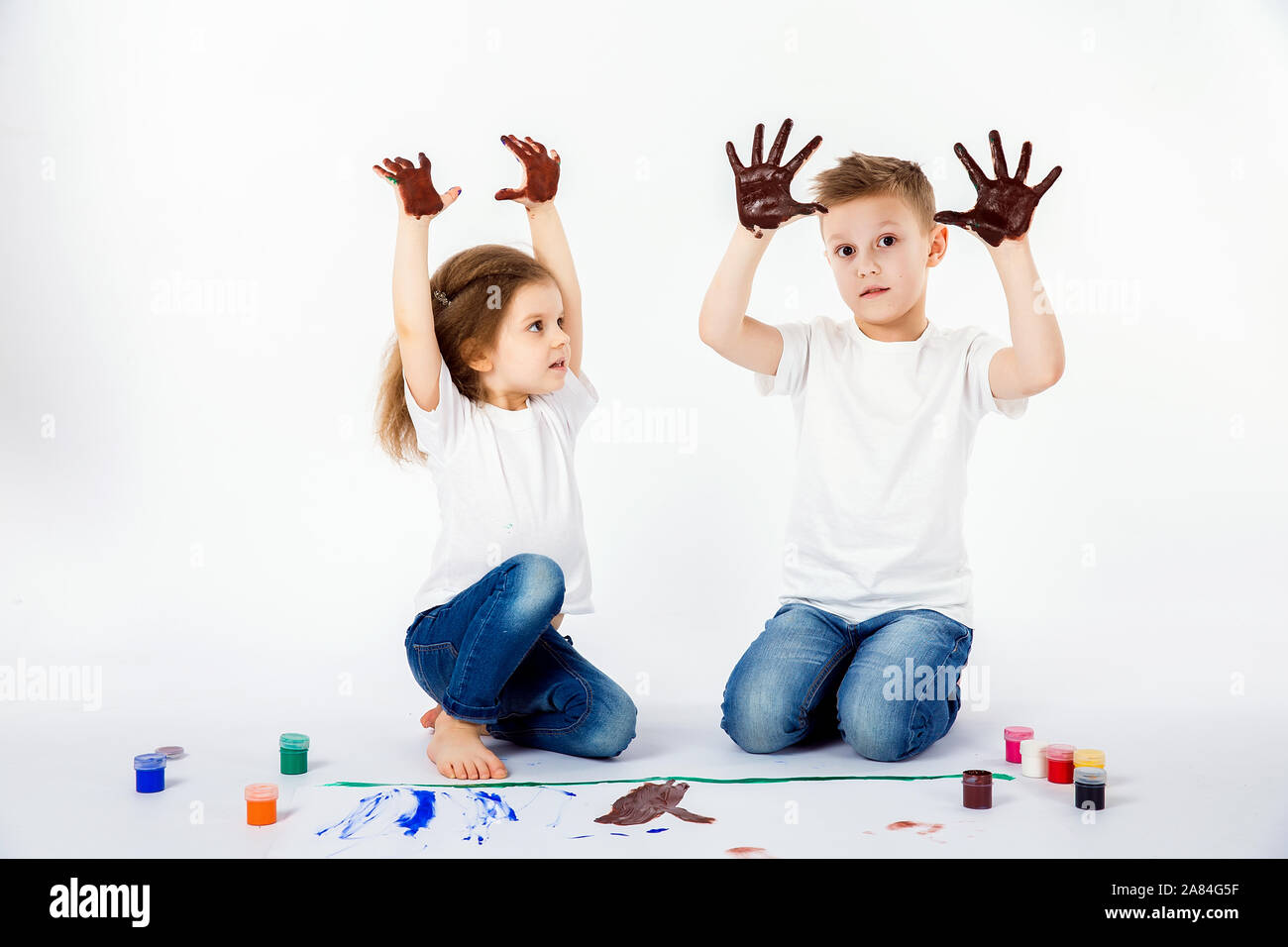 Two pretty child friends boy and girl in white shirts and blue jeans,  trendy hair style, barefoot, drawing pictures on white sheet of paper by  paints isolated on white. Showing hands in