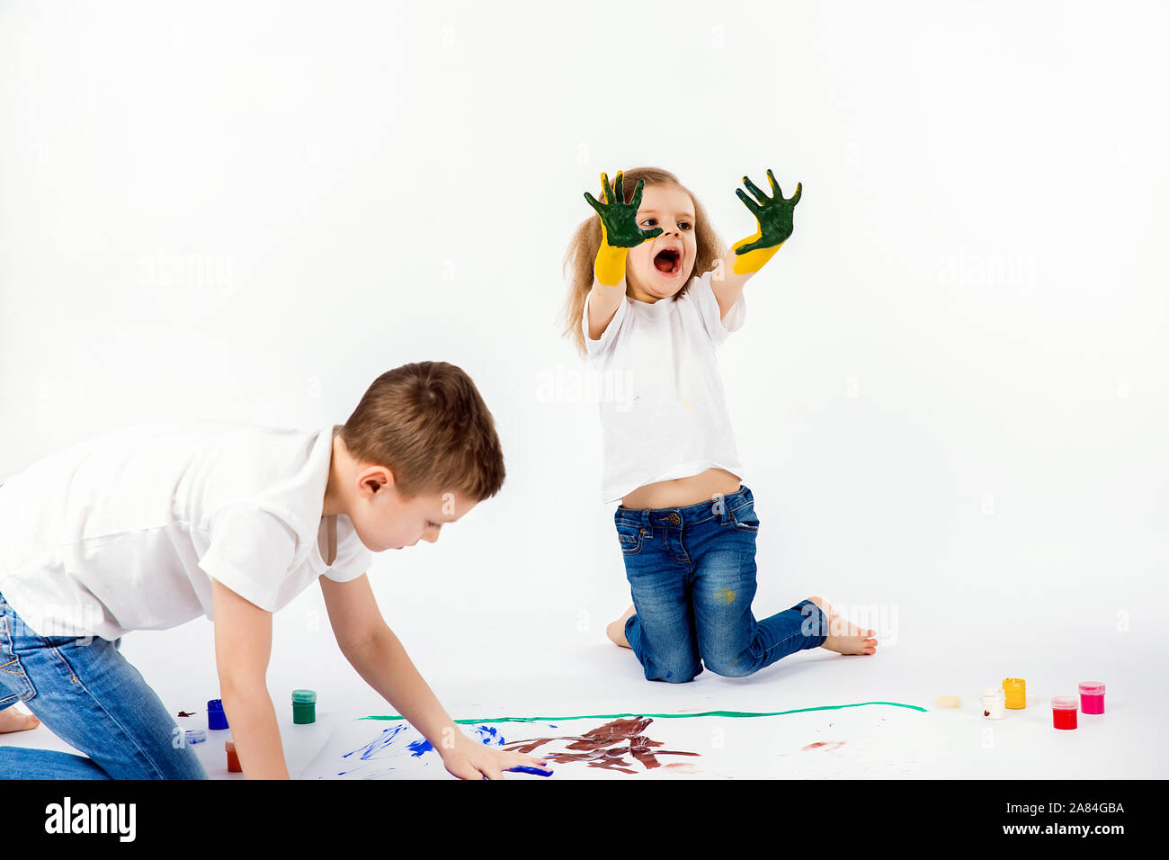 Two Pretty Child Friends Boy And Girl In White Shirts And Blue Jeans Trendy Hair Style Barefoot Drawing Pictures On White Sheet Of Paper By Paints Isolated On White Showing Hands In