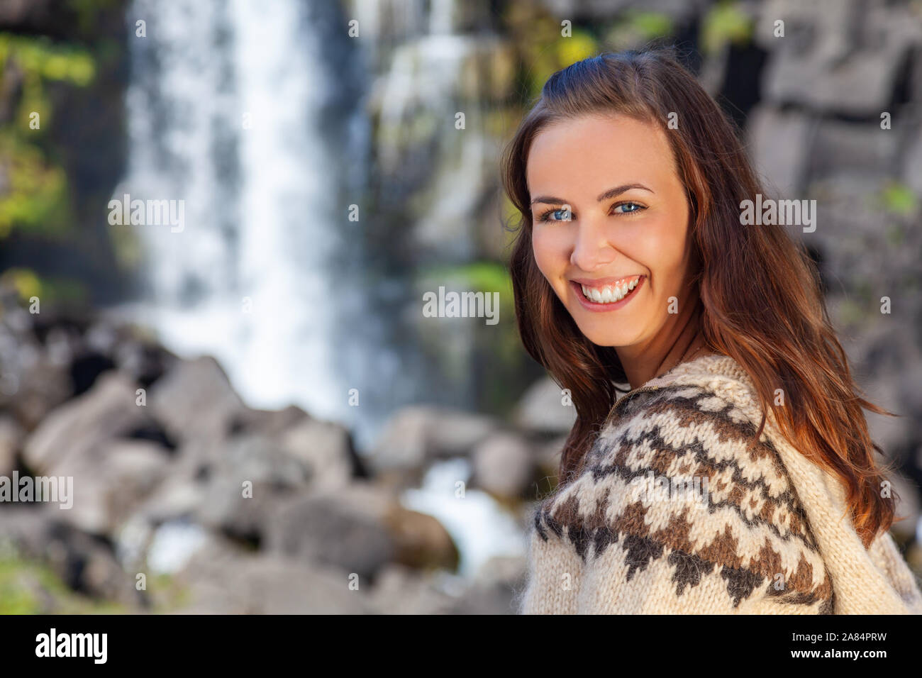 Beautiful Icelandic woman wearing traditionally patterned knitwear smiling in the mountains by a waterfall in Northern Iceland Stock Photo