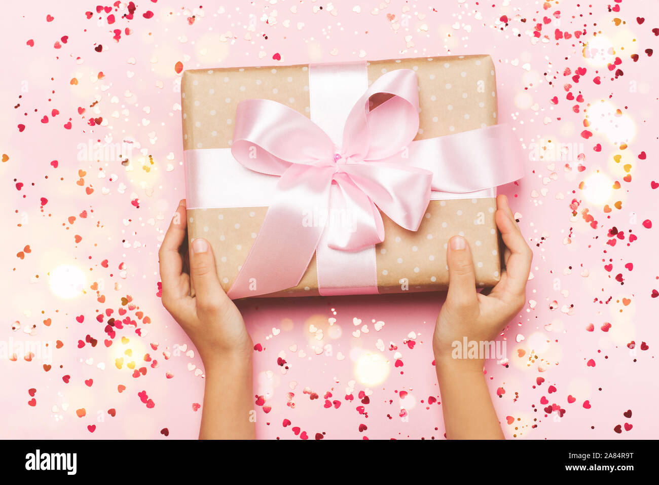 Girl's hands holding gift in kraft box with lush ribbon. Pink colors. Present for St. Valentine's day, Mother's Day, birthday, New Year, Christmas, holidays. Flat lay Stock Photo