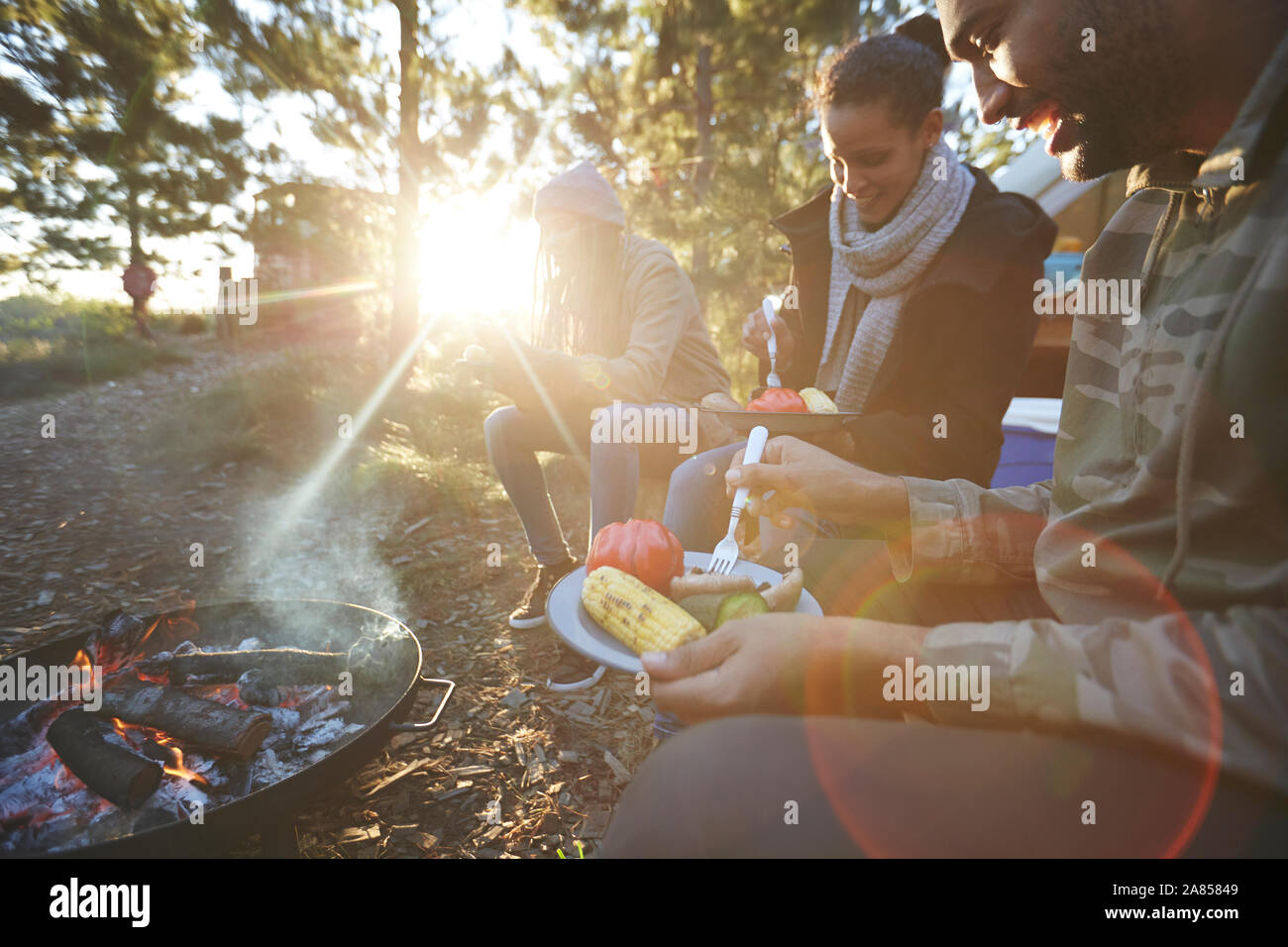 Family eating at sunny campsite campfire in woods Stock Photo