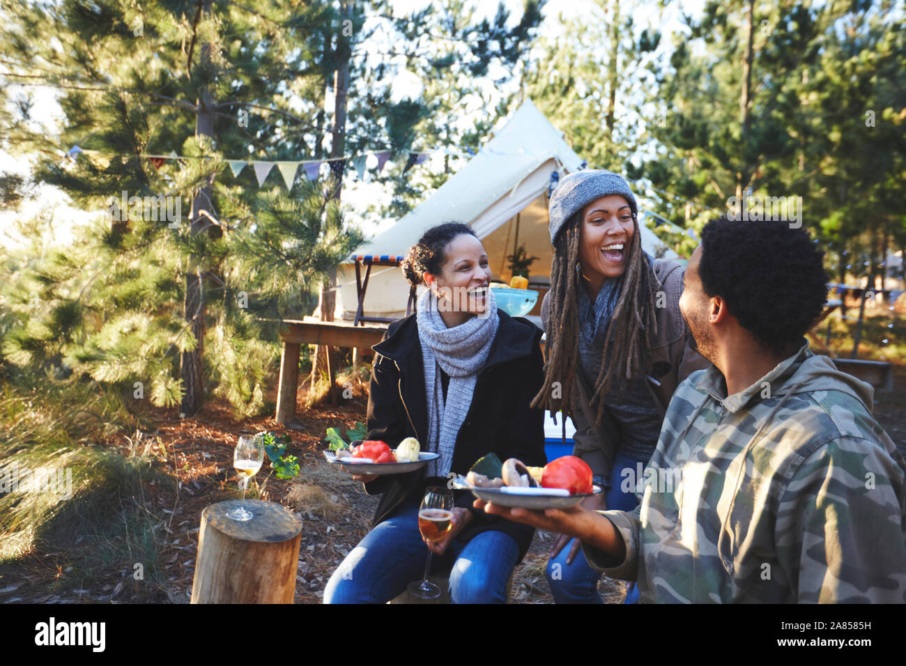 Happy friends laughing and eating at campsite in woods Stock Photo