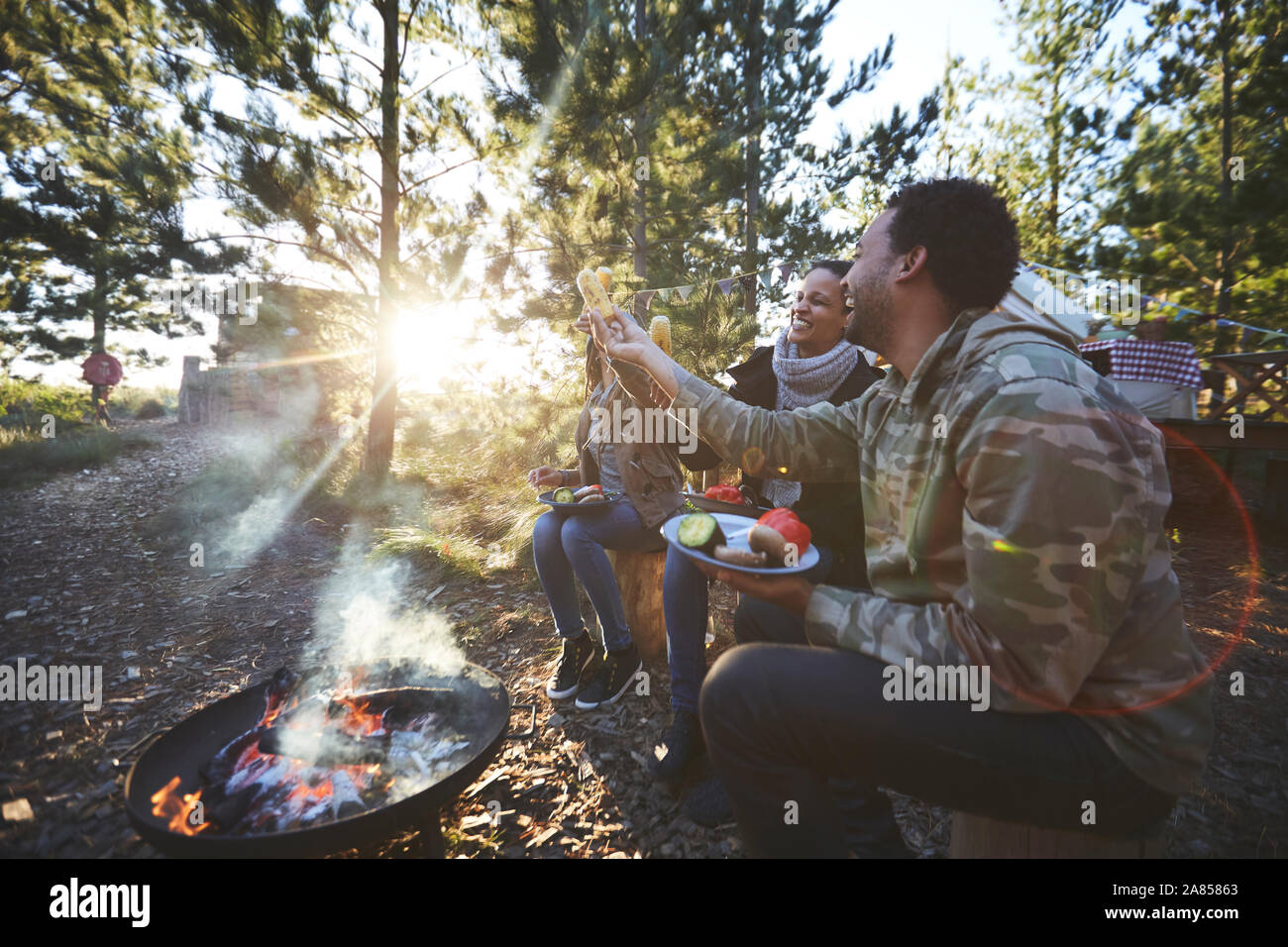 Happy friends eating at sunny campsite in woods Stock Photo
