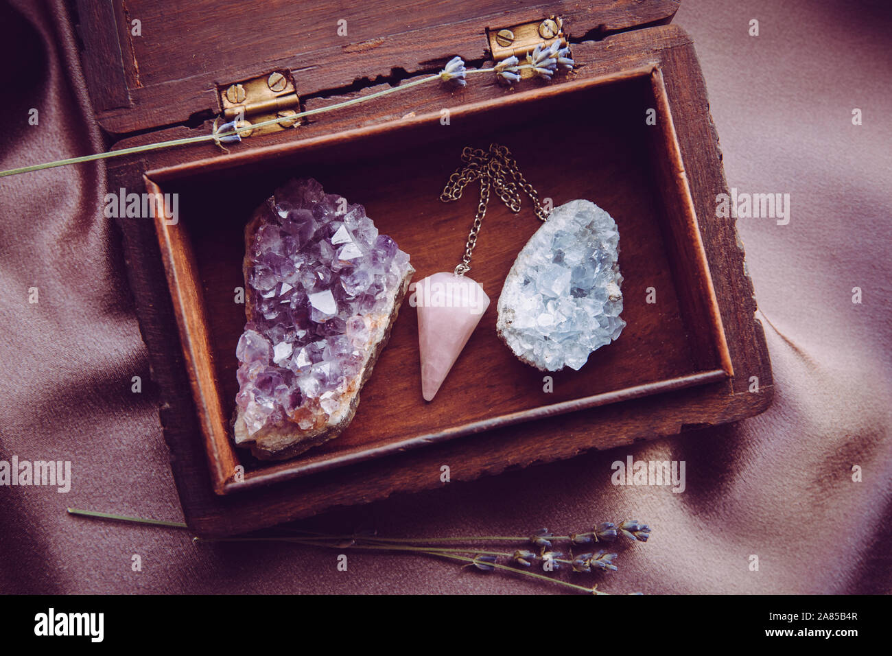 Witch tools inside beautiful old wood box. Rose quartz pendulum, natural amethyst and celestite crystal clusters. Dry lavender flowers on dark purple Stock Photo