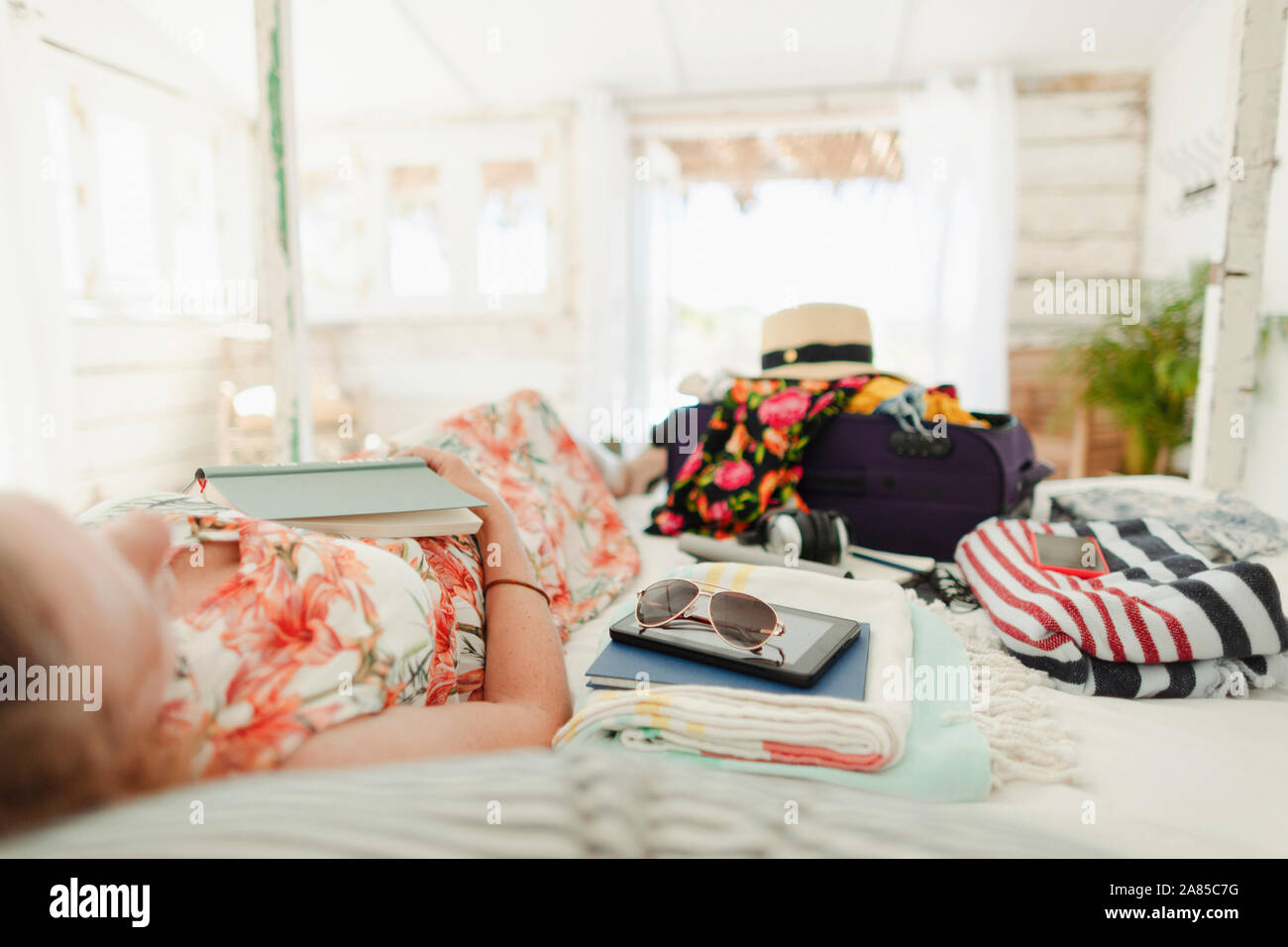 Woman with book relaxing on bed next to suitcase in sunny beach hut bedroom Stock Photo