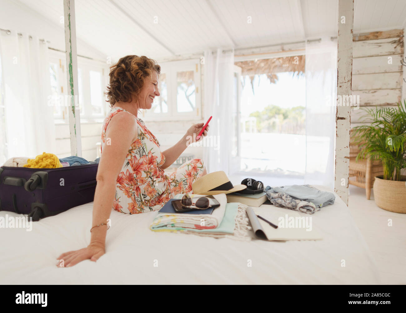 Woman with smart phone unpacking suitcase on beach hut bed Stock Photo