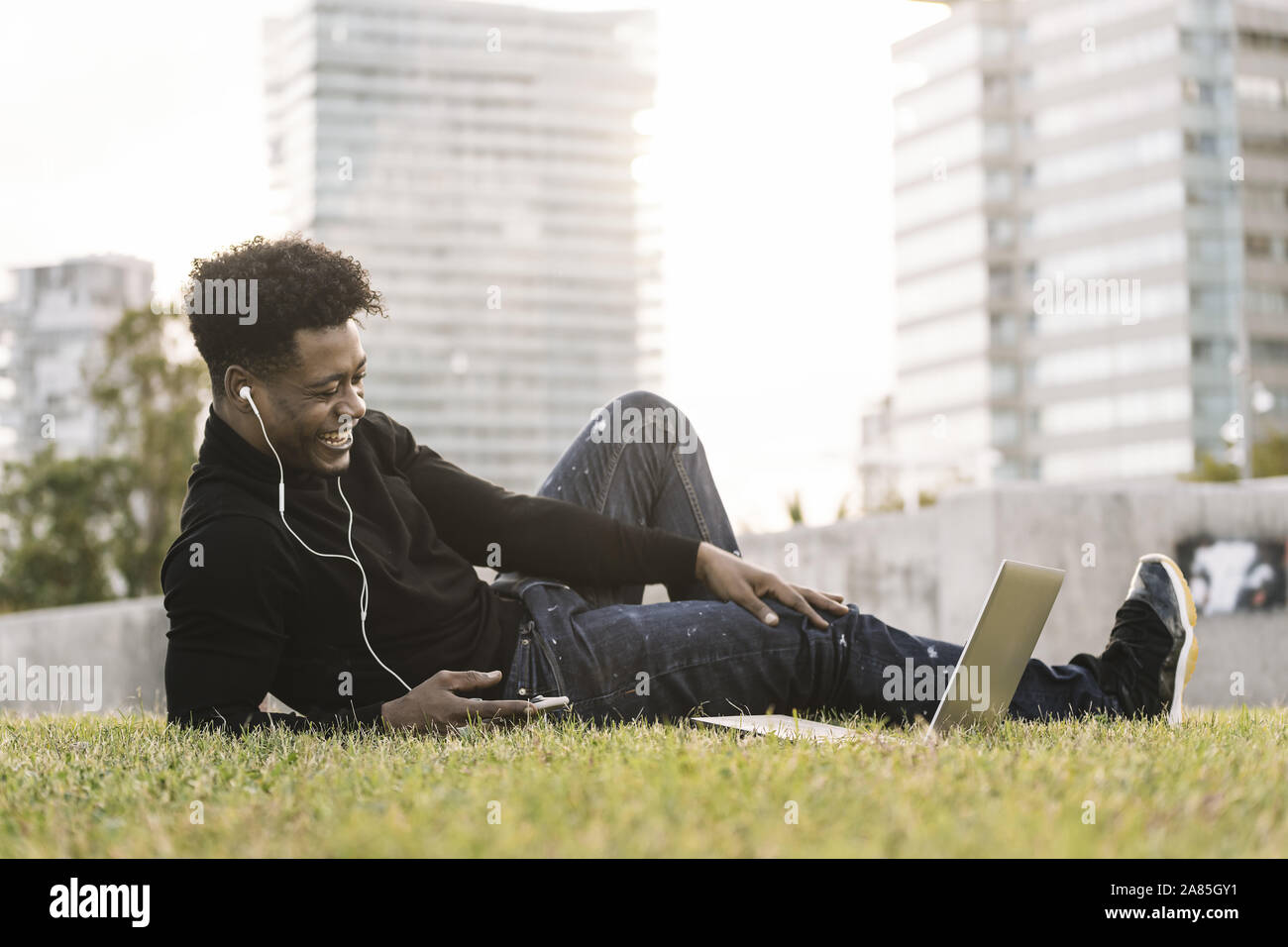 smiling young black man laughing with white earphones looking to the laptop lying on the lawn in the city at sunset, lifestyle and technology concept Stock Photo