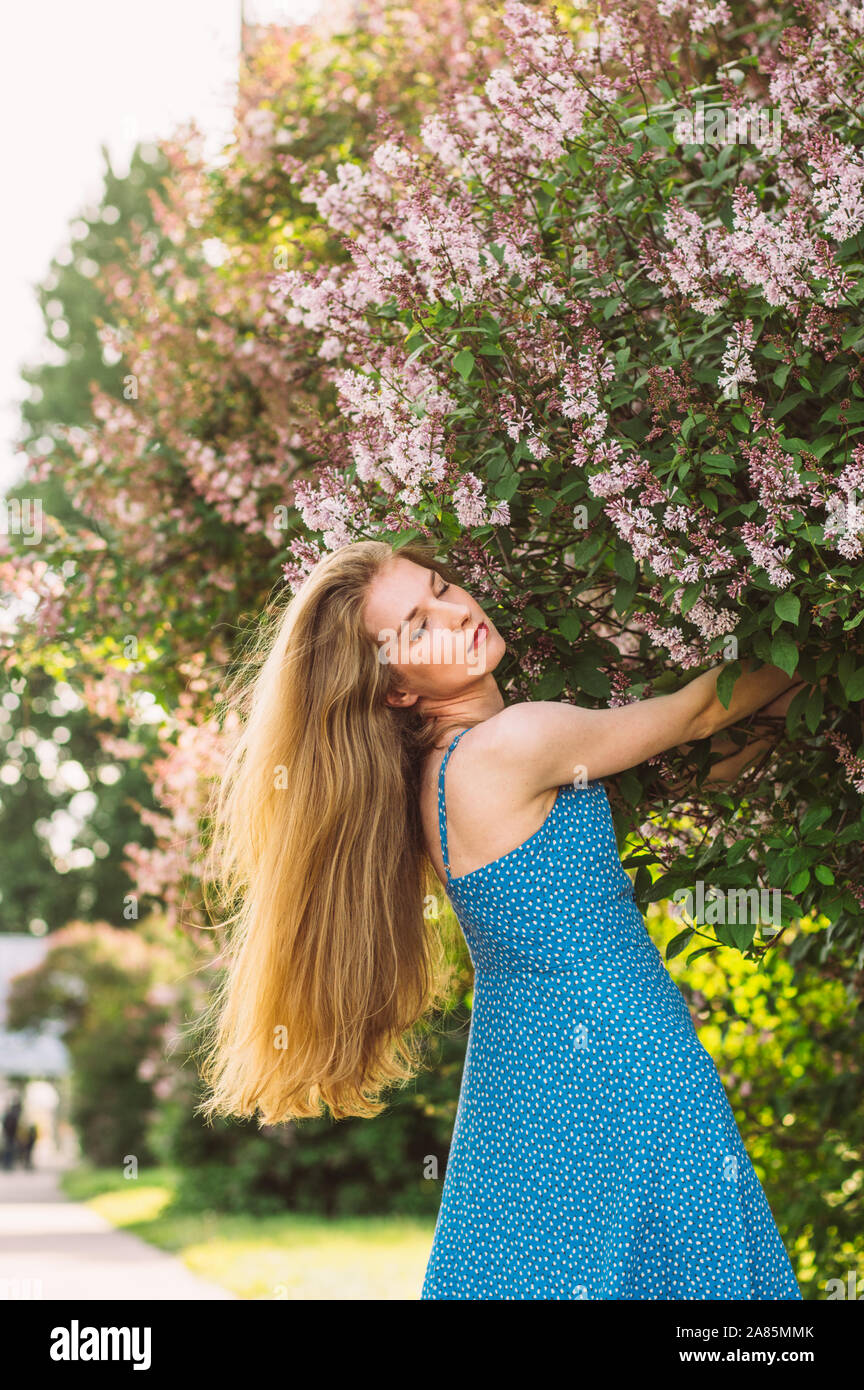 Portrait of girl with blond hair and closed eyes and blue dress among lilac, summertime Stock Photo
