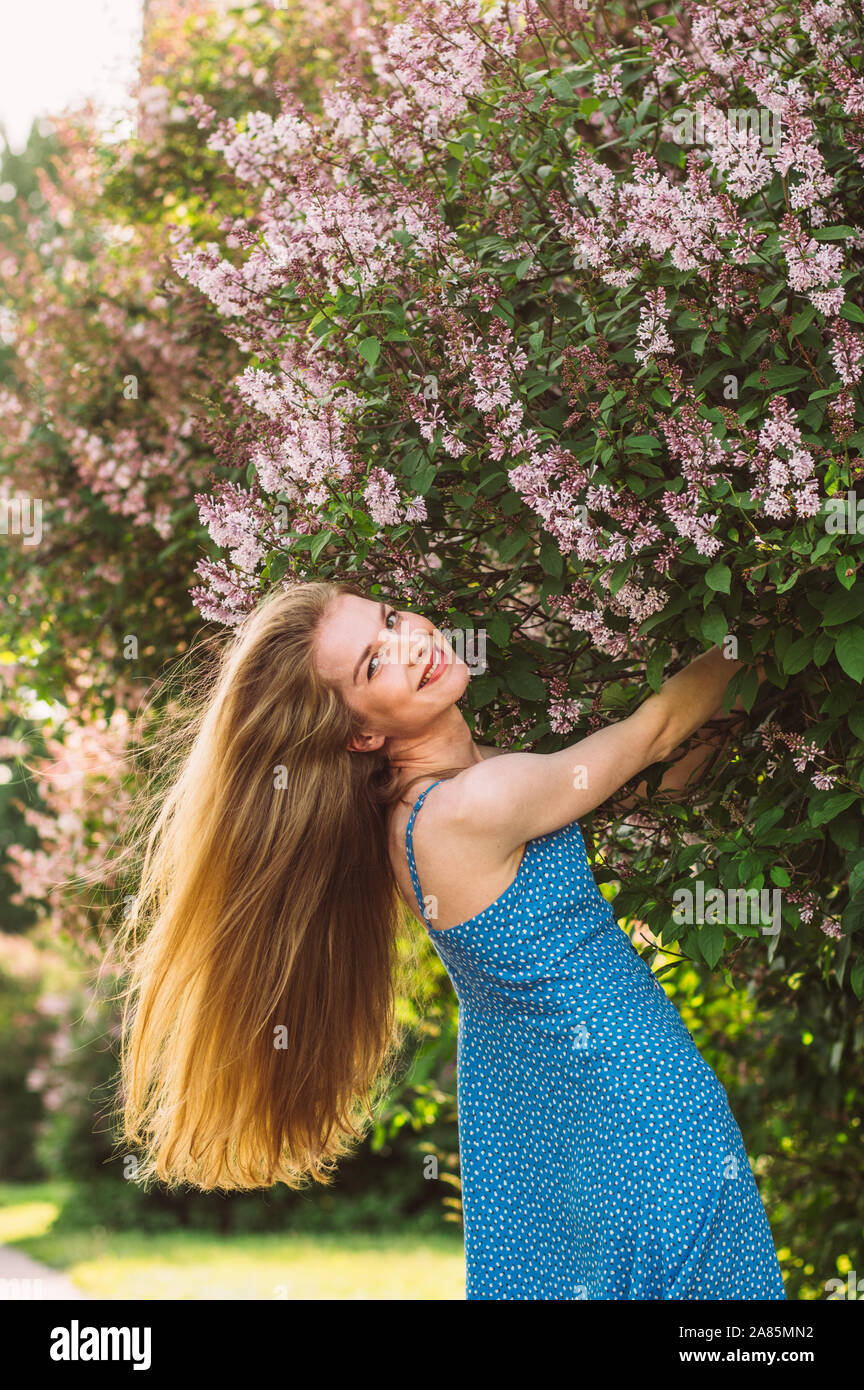 Portrait of smiling girl with blond hair and blue dress among lilac, summertime Stock Photo