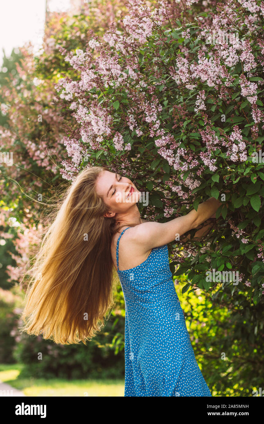 Portrait of smiling girl with blond hair and closed eyes and blue dress among lilac, summertime Stock Photo
