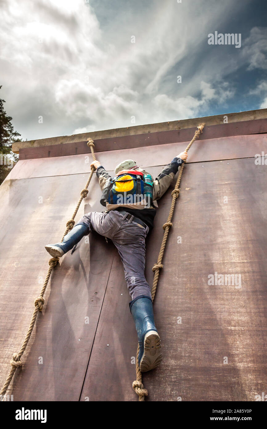 Young boy in wellys climbing up a wall with ropes. Stock Photo