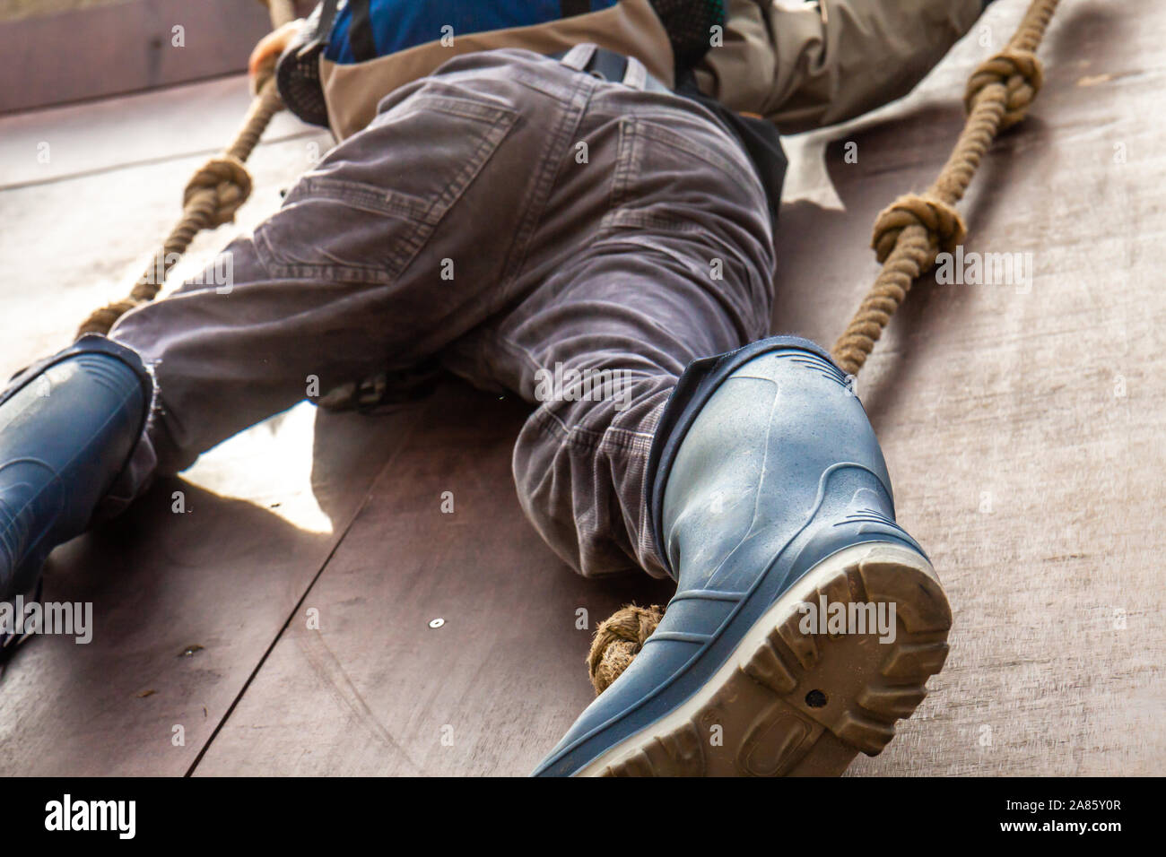Young boy in wellys climbing up a wall with ropes. Stock Photo
