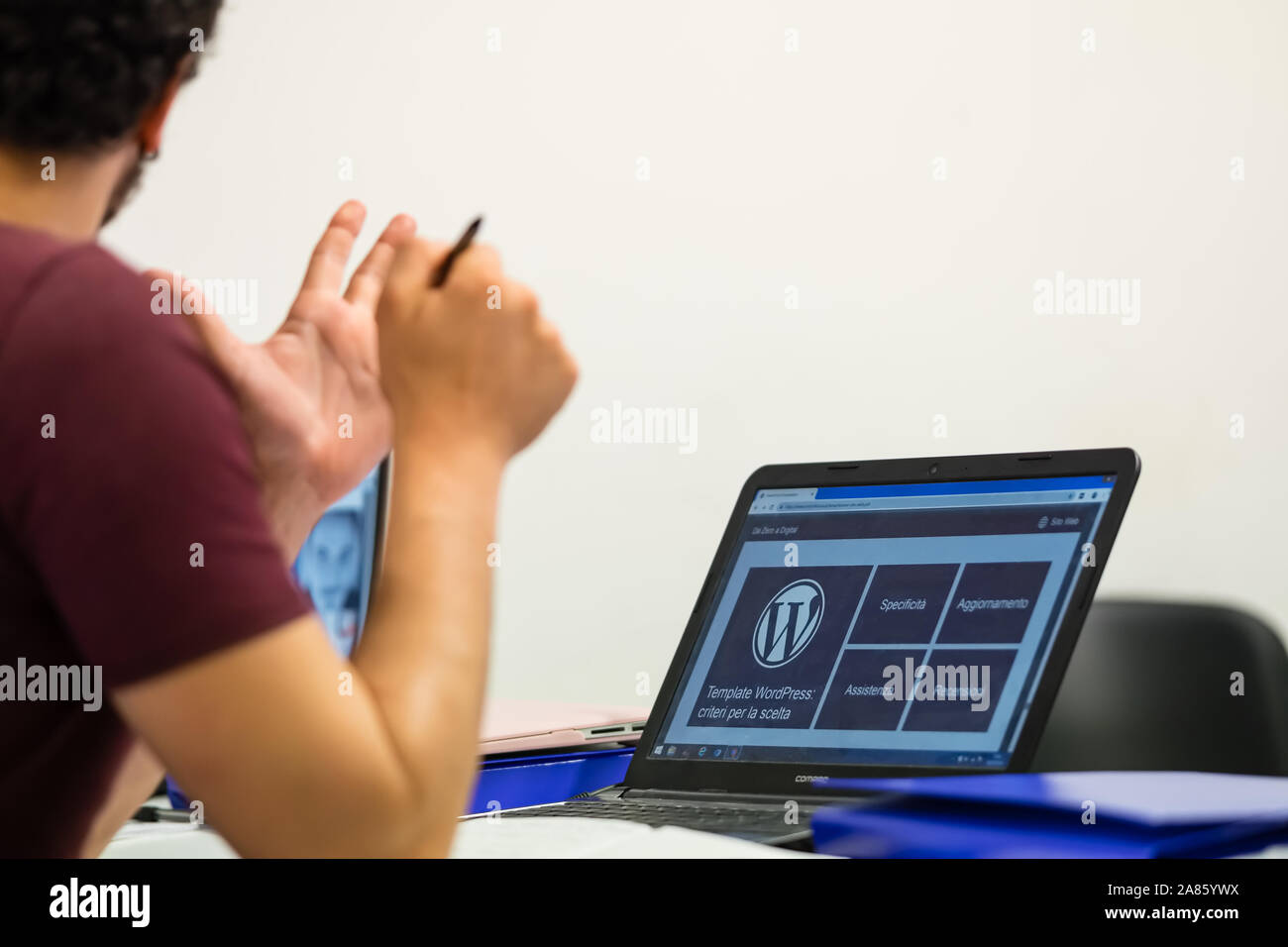 Laptop photographed with the Wordpress logo on the screen, young operator partially framed from behind with a pen in his hand. Stock Photo