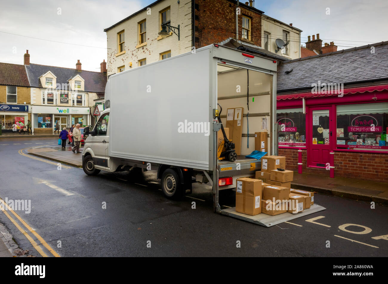 A white van delivering goods to a chocolate shop with boxed good lowered to ground on a tail lift. Stock Photo
