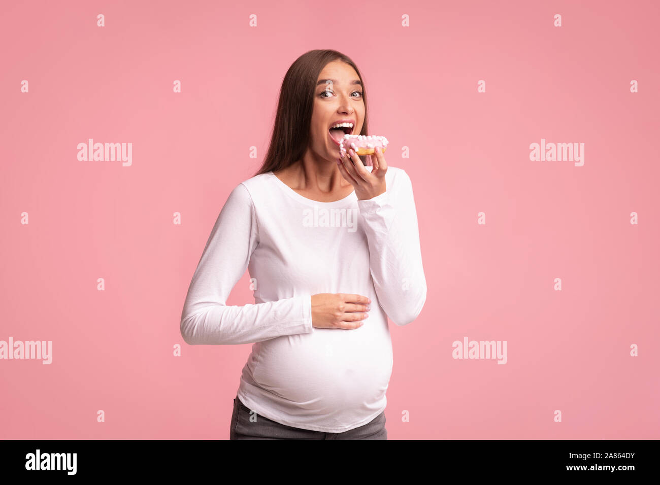 Pregnant Woman Eating Donut On Pink Background, Studio Shot Stock Photo