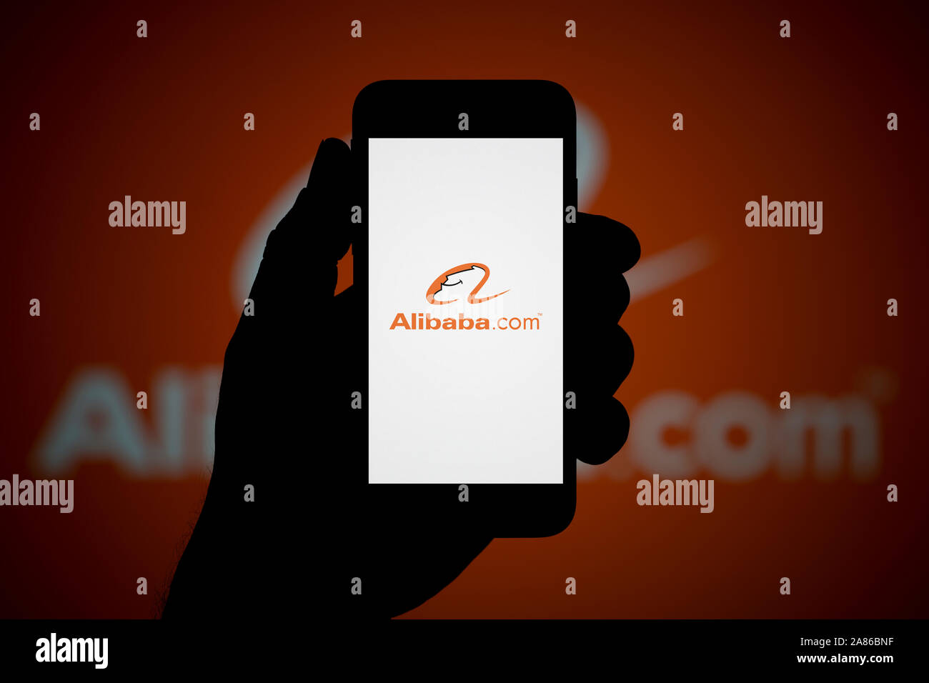 A man looks at his iPhone which displays the Alibaba logo, with the same logo in the background (Editorial use only). Stock Photo