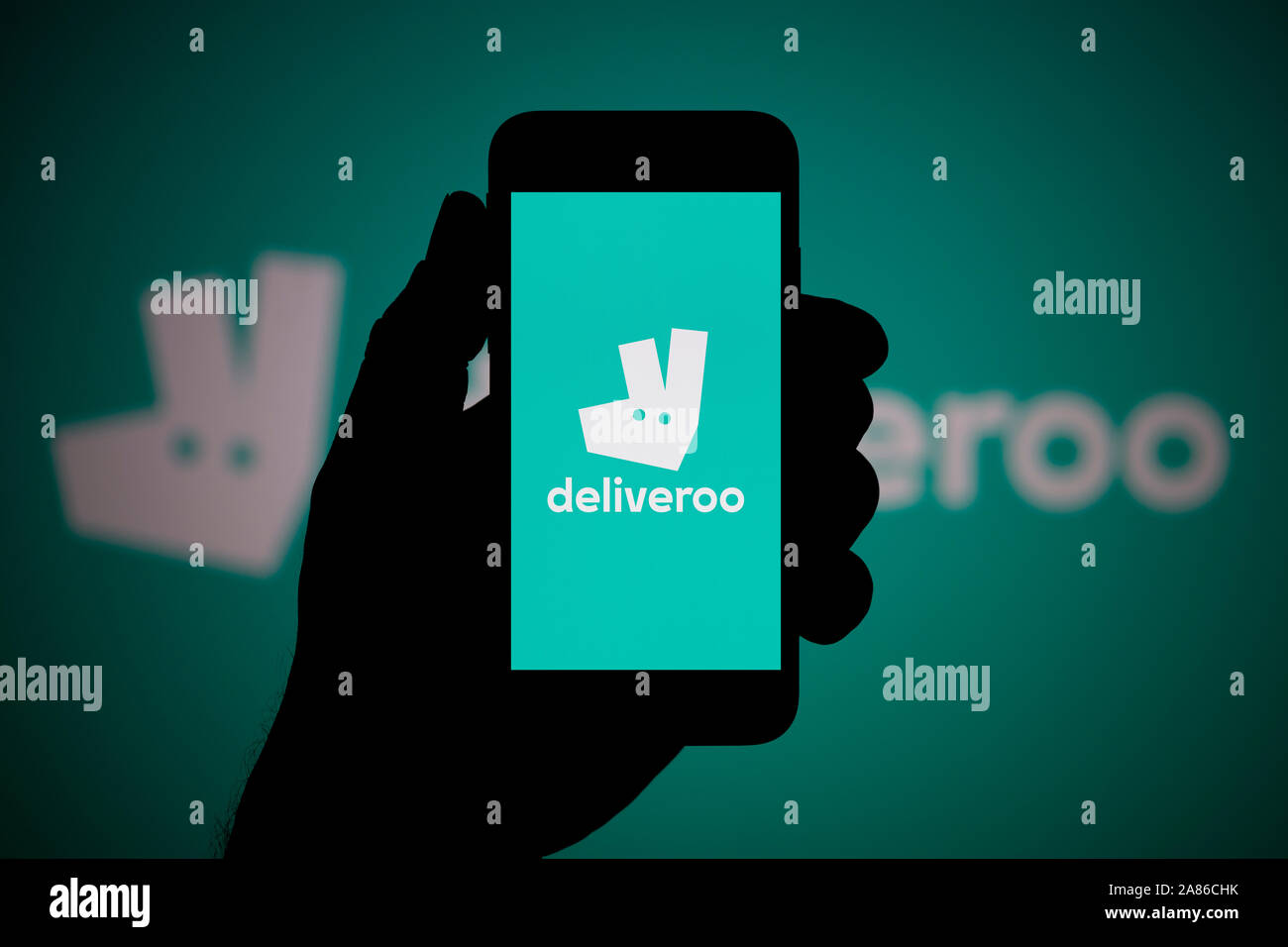 A man looks at his iPhone which displays the Deliveroo logo, with the same logo in the background (Editorial use only). Stock Photo