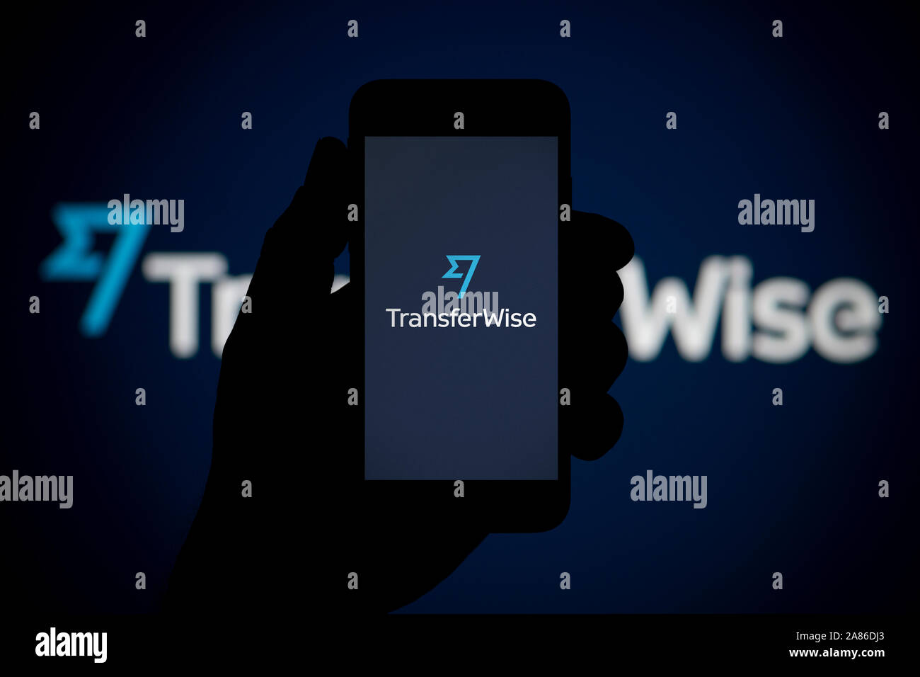 A man looks at his iPhone which displays the TransferWise logo, with the same logo in the background (Editorial use only). Stock Photo
