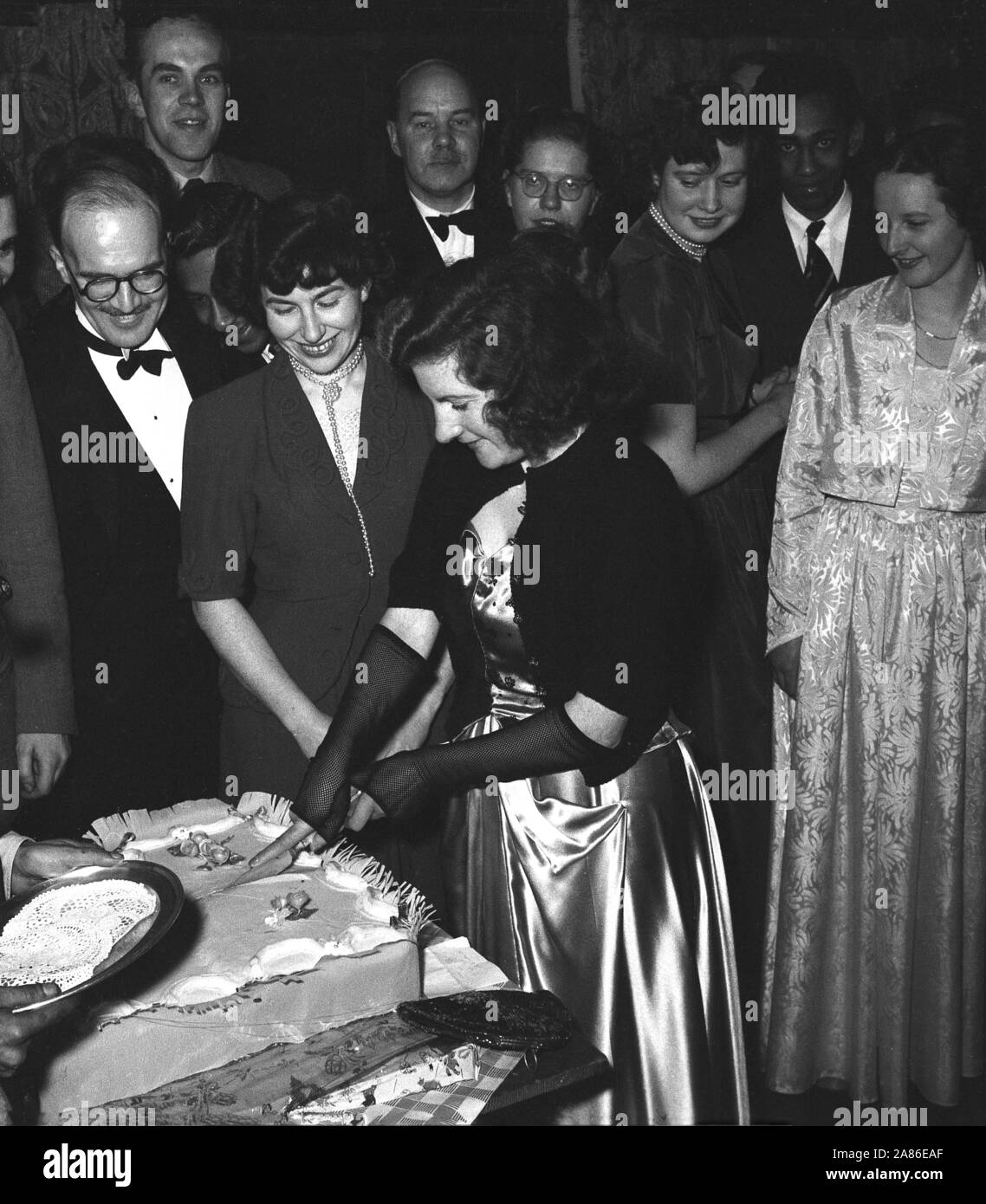 1950s, historical, adult guests in formal attire at a black tie birthday party, watch as a young lady wearing a shiny long dress, a shawl covering her shoulders and black lace armlets, uses both of her hands to cut a large cake. A metal dish awaits the first slice. Stock Photo