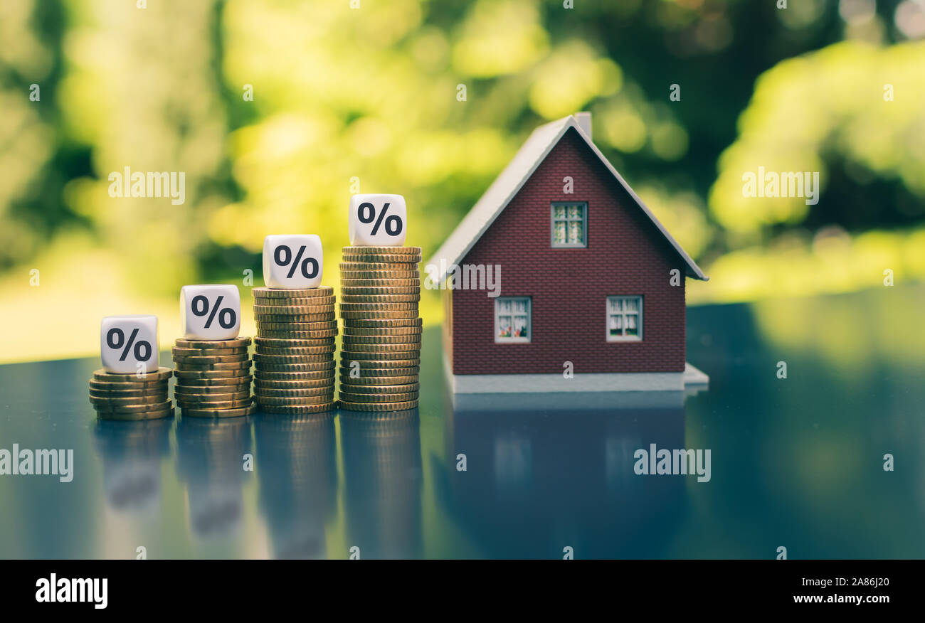Symbol for increasing interest rates. Dice with percentage symbols on increasing high stacks of coins next to a model house. Stock Photo
