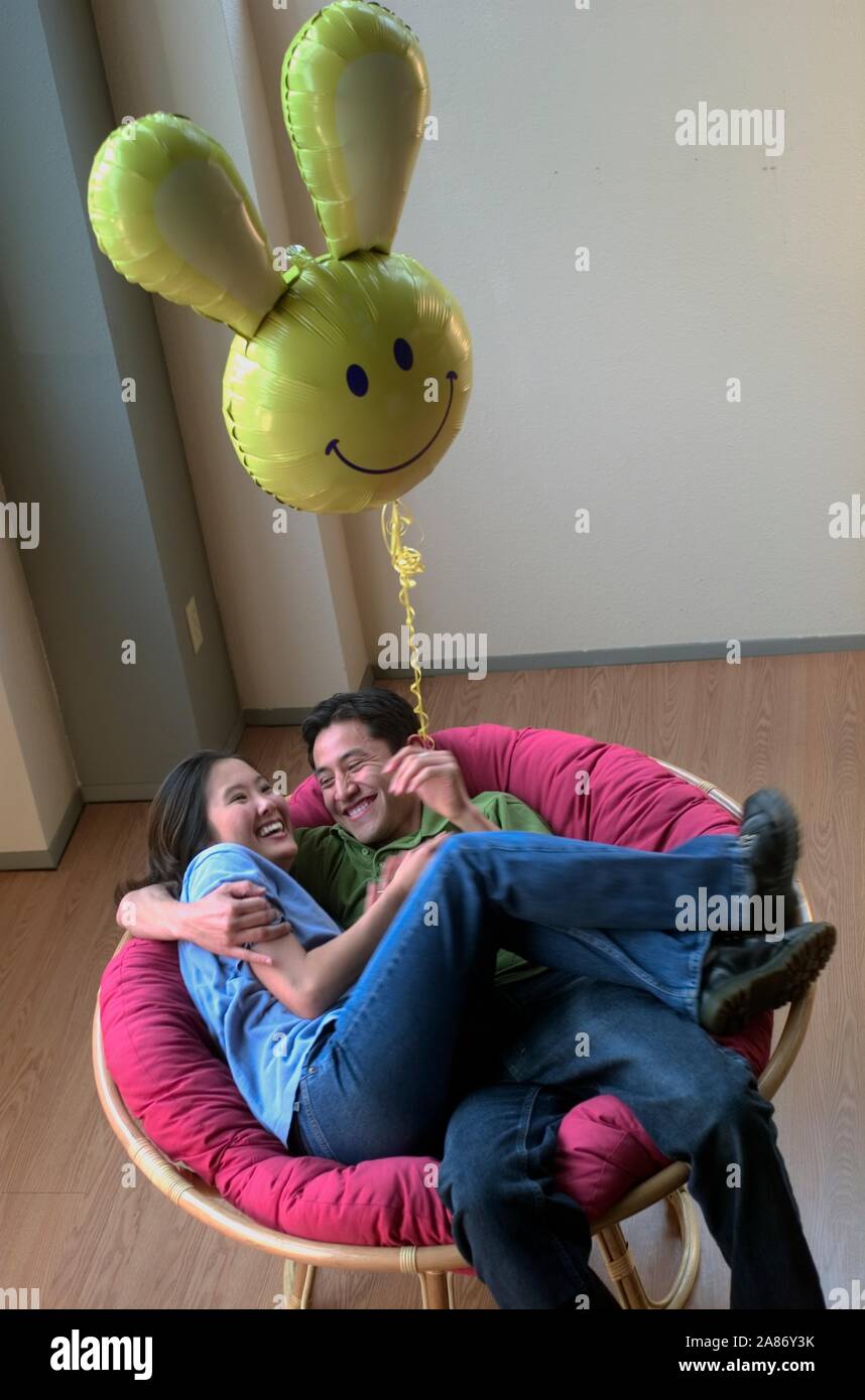 Asian American couple hanging out together in a big chair with a silly balloon hovering above Stock Photo