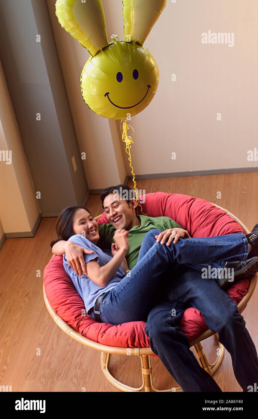 Asian American couple hanging out together in a big chair with a silly balloon hovering above Stock Photo