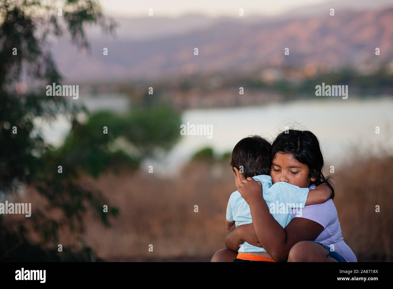 Brother and sister separated from parents are hugging each other in a foreign country, across a river. Stock Photo