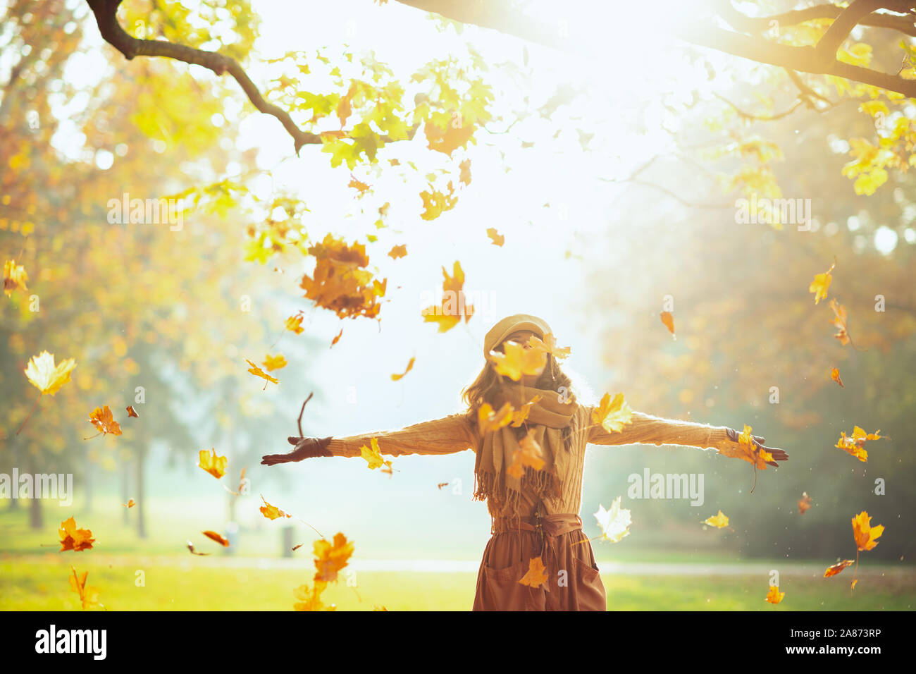 Hello autumn. elegant woman in sweater, skirt, hat, gloves and scarf outdoors in the autumn park enjoying autumn and catching falling yellow leaves. Stock Photo