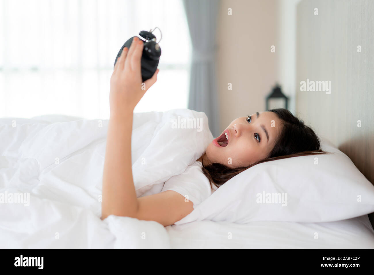 Beautyful Asian Woman lying on white bed and holding black alarm clock with shock to learn that late in morning at bedroom at home. Stock Photo