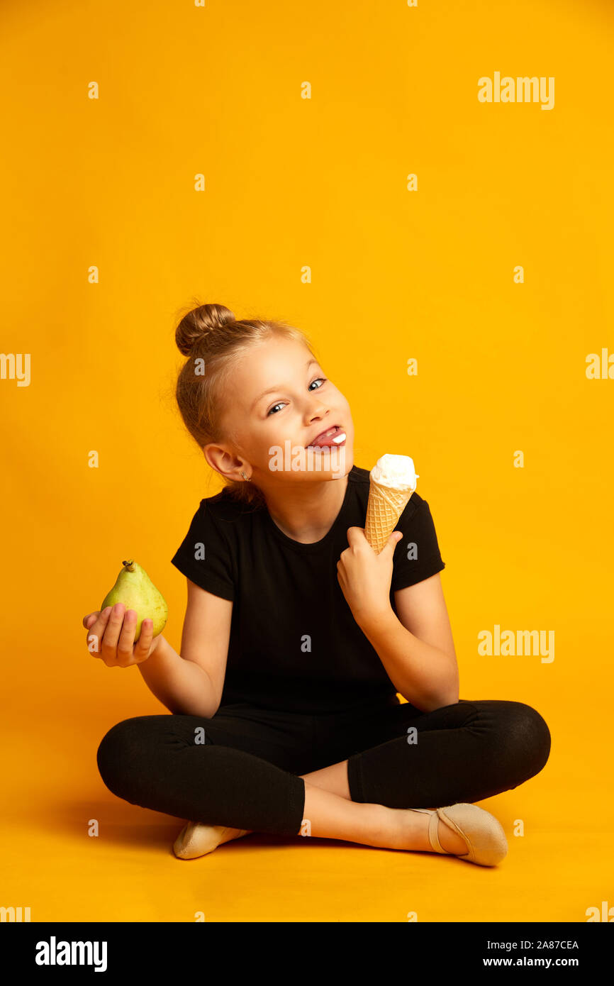 Cute little gymnast girl choosing between pear and sweet ice-cream on yellow background Stock Photo