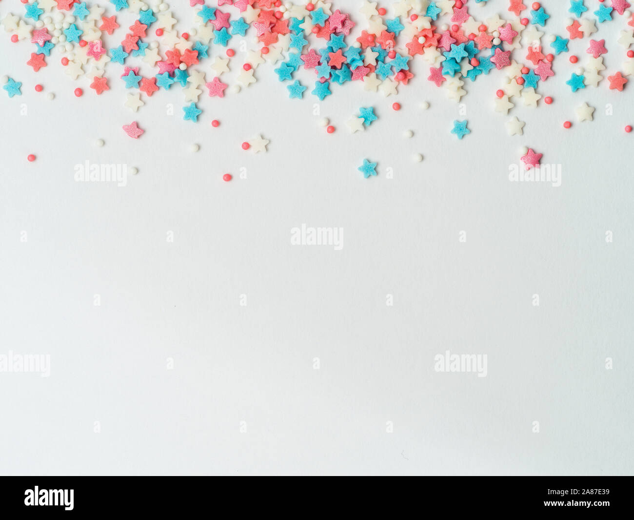 Festive border frame of colorful pastel sprinkles on white background, copy space bottom. Sugar sprinkle dots and stars, decoration for cake and bakery. Top view or flat lay Stock Photo