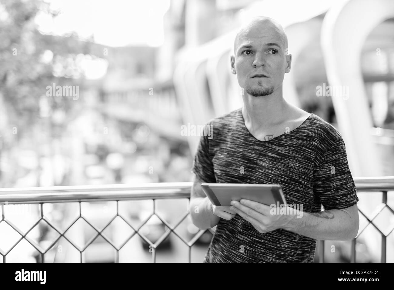 Young handsome bald man thinking while holding digital tablet on the footbridge against view of the city streets Stock Photo