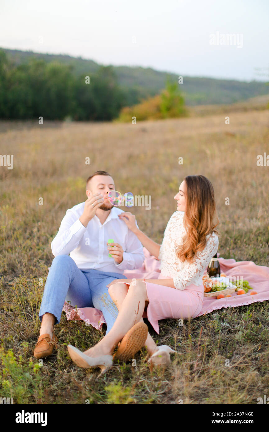 Young jocund man blowing soap bubbles near caucasian girl and sitting on plaid in steppe. Stock Photo