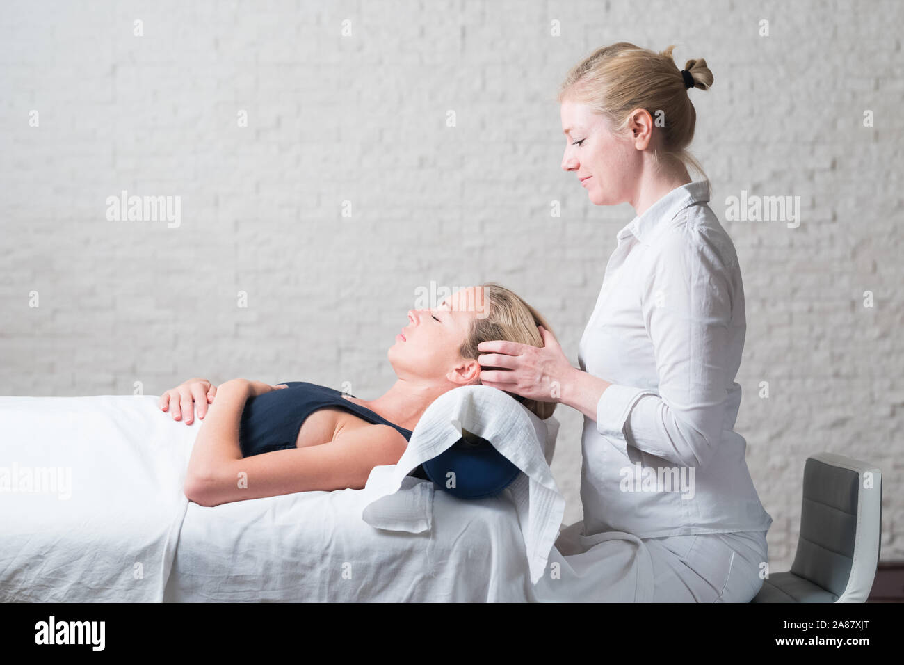 Professional female masseur giving relaxing massage treatment to young female client. Hands of masseuse on forehead of young lady during procedure of Stock Photo