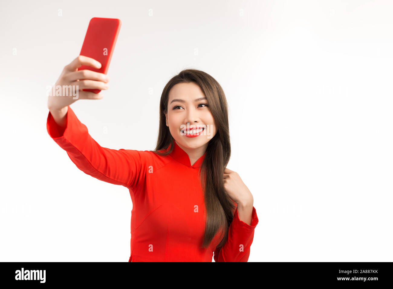 Asian woman in Vietnamese dress traditional ao dai. Beautiful toothy smiling young woman using smartphone doing FaceTime with friends against white ba Stock Photo