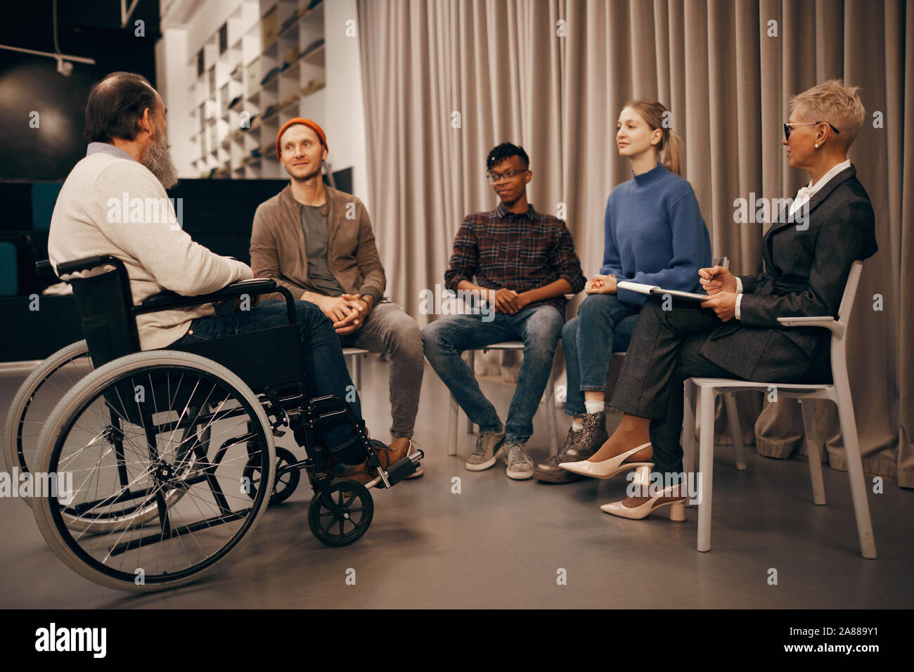 Group of business people sitting on chairs and listening to senior man who sitting in wheelchair at business meeting Stock Photo