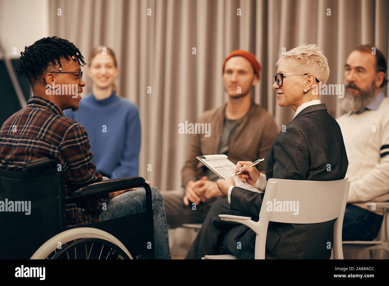 Young African man in wheelchair talking to mature woman while she making notes during therapy lesson Stock Photo