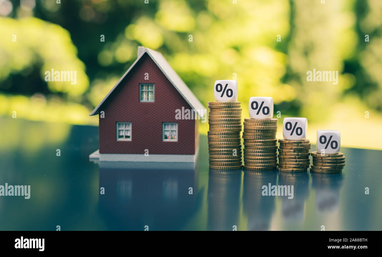 Symbol for decreasing interest rates. Dice with percentage symbols on decreasing high stacks of coins next to a model house. Stock Photo