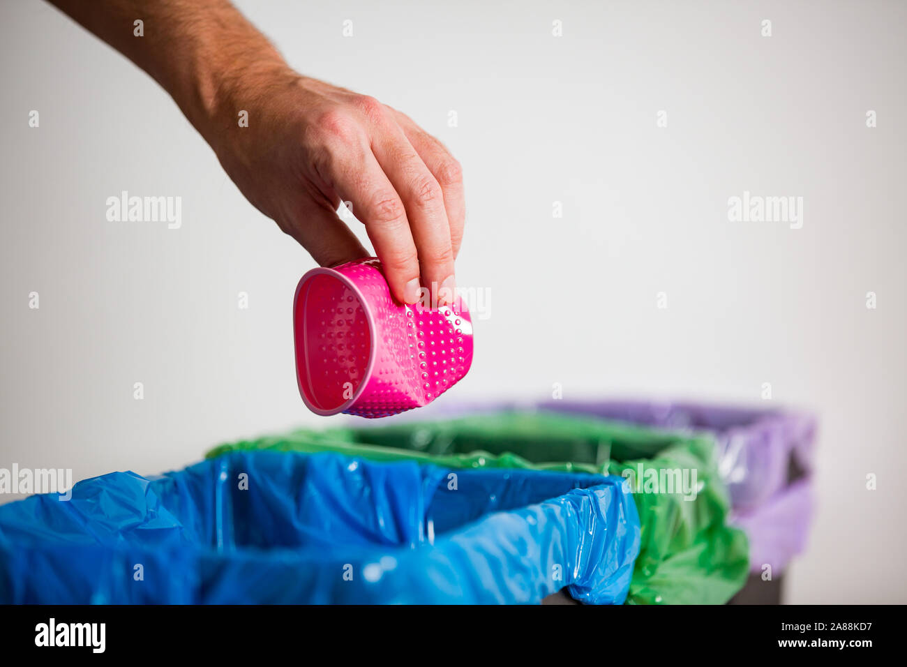 Hand putting single-use plastic cup into recycling bin. Person in a house kitchen separating waste. Black trash bin with green bag and recycling Stock Photo